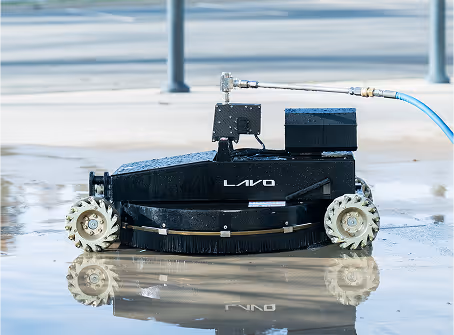 Robotic floor cleaner with bristled brush, labeled "Lavo," on wet concrete. Industrial setting, connected to a blue hose, reflecting light.