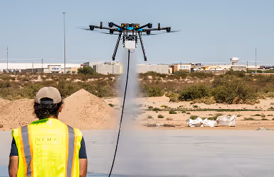 A person in a neon vest operates a large spraying drone over a construction site, with sandy terrain and industrial buildings in the background.