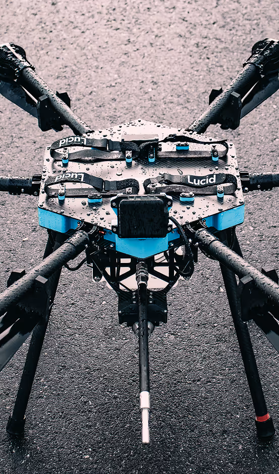 A large drone with visible propeller arms is set on a wet pavement. Its blue and black frame is covered with raindrops, conveying a technical and rugged tone.