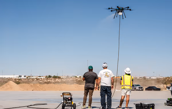 Three people observe a flying drone spraying water in an open, dry landscape. They stand on pavement, wearing casual and work attire under a clear sky.
