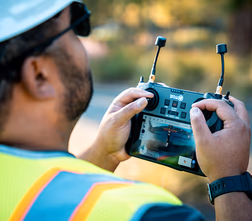 A person in a safety vest and helmet operates a remote drone controller, focusing intently on the screen. The setting is outdoors, suggesting drone use.