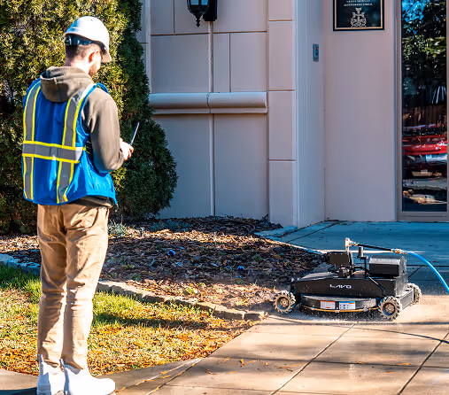 A worker wearing a hard hat and blue vest operates a remote-controlled robot on a concrete pathway. The robot is inspecting or cleaning the path.