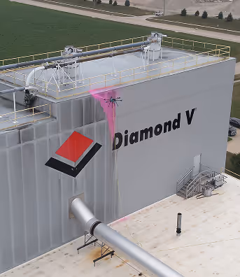 A drone sprays pink liquid on the side of a large industrial building marked 'Diamond V.' A vast green field is visible in the background, conveying a sense of innovation.