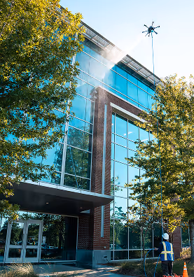 A worker uses a tall pole to clean large glass windows on a modern building. Water sprays from the top, with trees framing the scene on a sunny day.