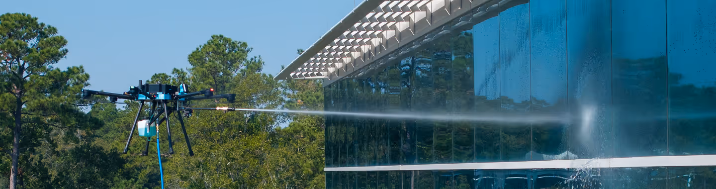 A drone sprays water on a glass building surrounded by trees. The scene conveys modern technology in maintenance amidst a natural setting.