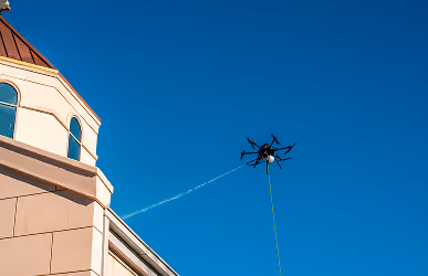 A drone with multiple propellers sprays water near a building's corner against a clear blue sky, suggesting maintenance or cleaning work.