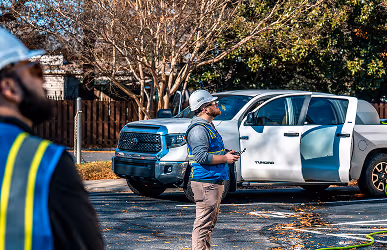 Workers in safety gear and helmets stand near a white pickup truck in a tree-lined area. The scene conveys an industrious and focused atmosphere.