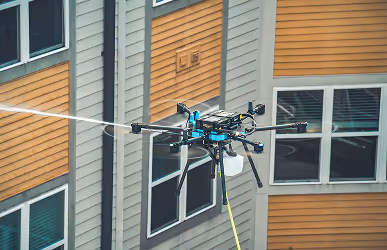 A drone sprays water while hovering near a building with orange and gray siding and multiple windows, suggesting exterior maintenance work.