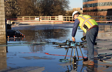 A construction worker in a safety vest and helmet operates a large drone on a sunny building site. Reflections shimmer on a wet concrete surface.