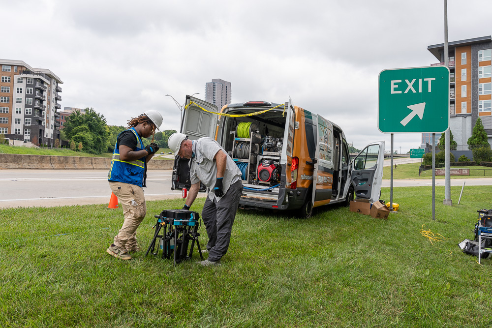 Workers in safety gear prepare drone equipment beside a service van near a highway exit.