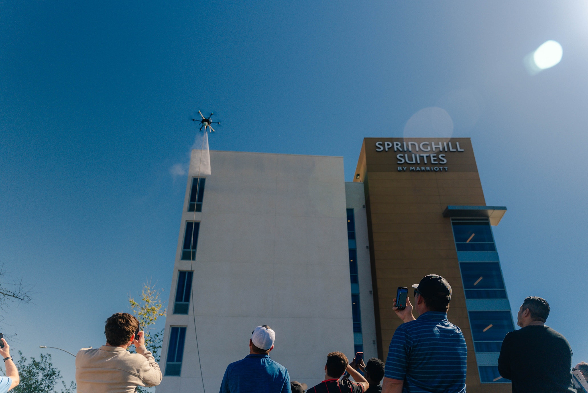 Drone cleaning the exterior facade of a Springhill Suites hotel while a crowd watches and films from the ground below