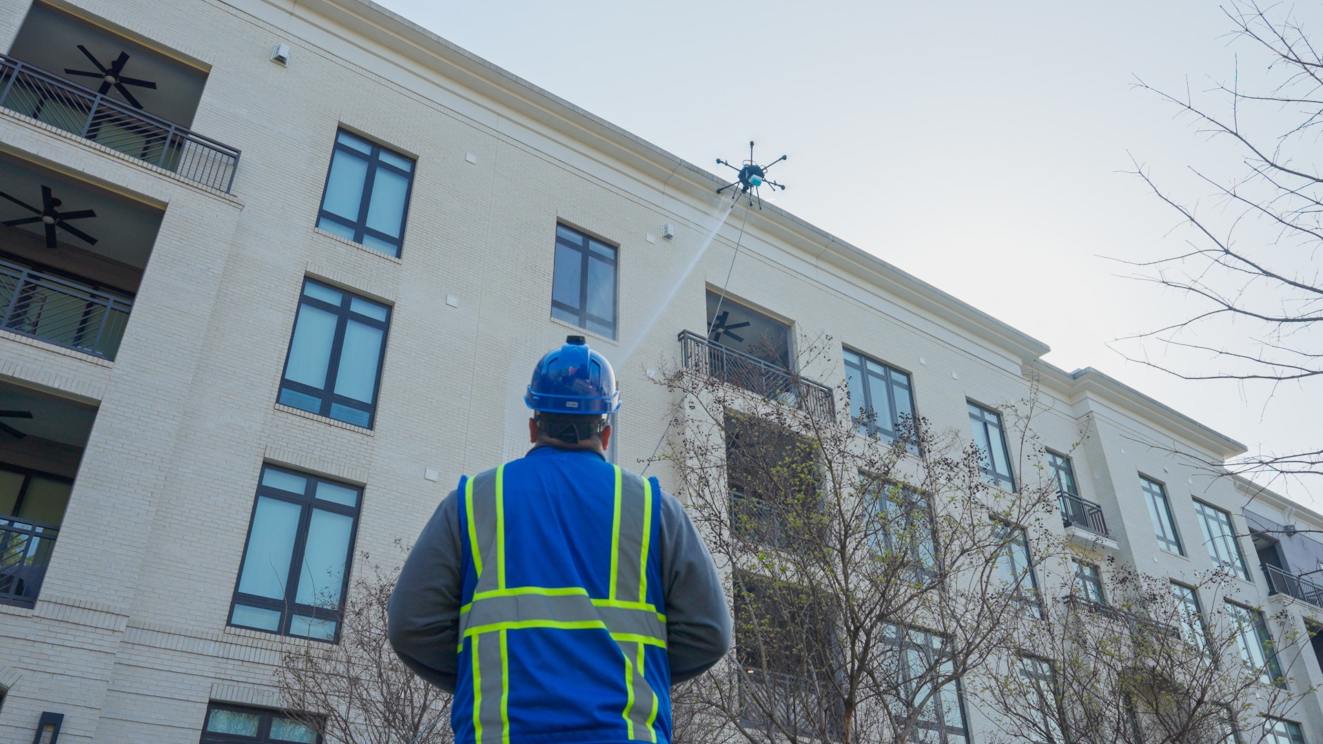Commercial drone delivering cleaning solution to the exterior facade of a multi-story building with a ground-based operator visible below