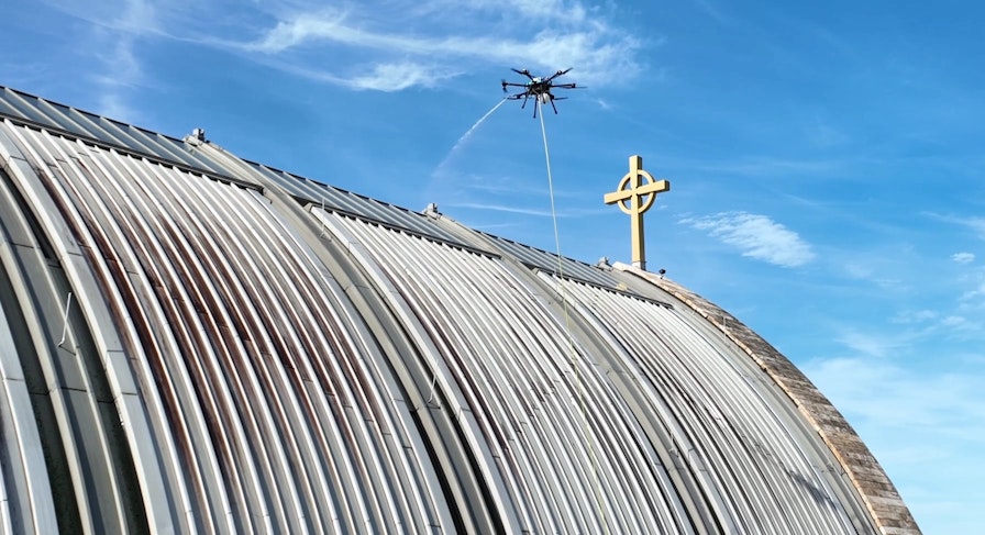 A Lucid Bots drone sprays water to clean the curved metal roof of the Ave Maria Oratory, with a gold Celtic cross visible against a blue sky.