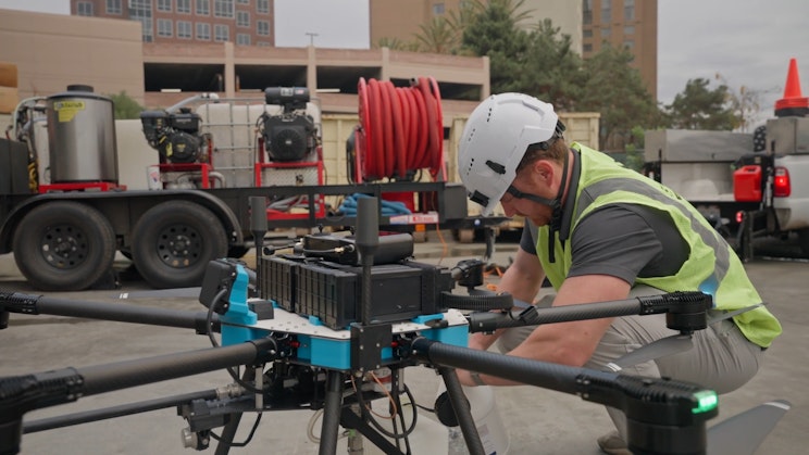 A technician in a safety vest and hard hat prepares a Lucid Bots drone for deployment, with pressure washing equipment on a trailer visible in the background.