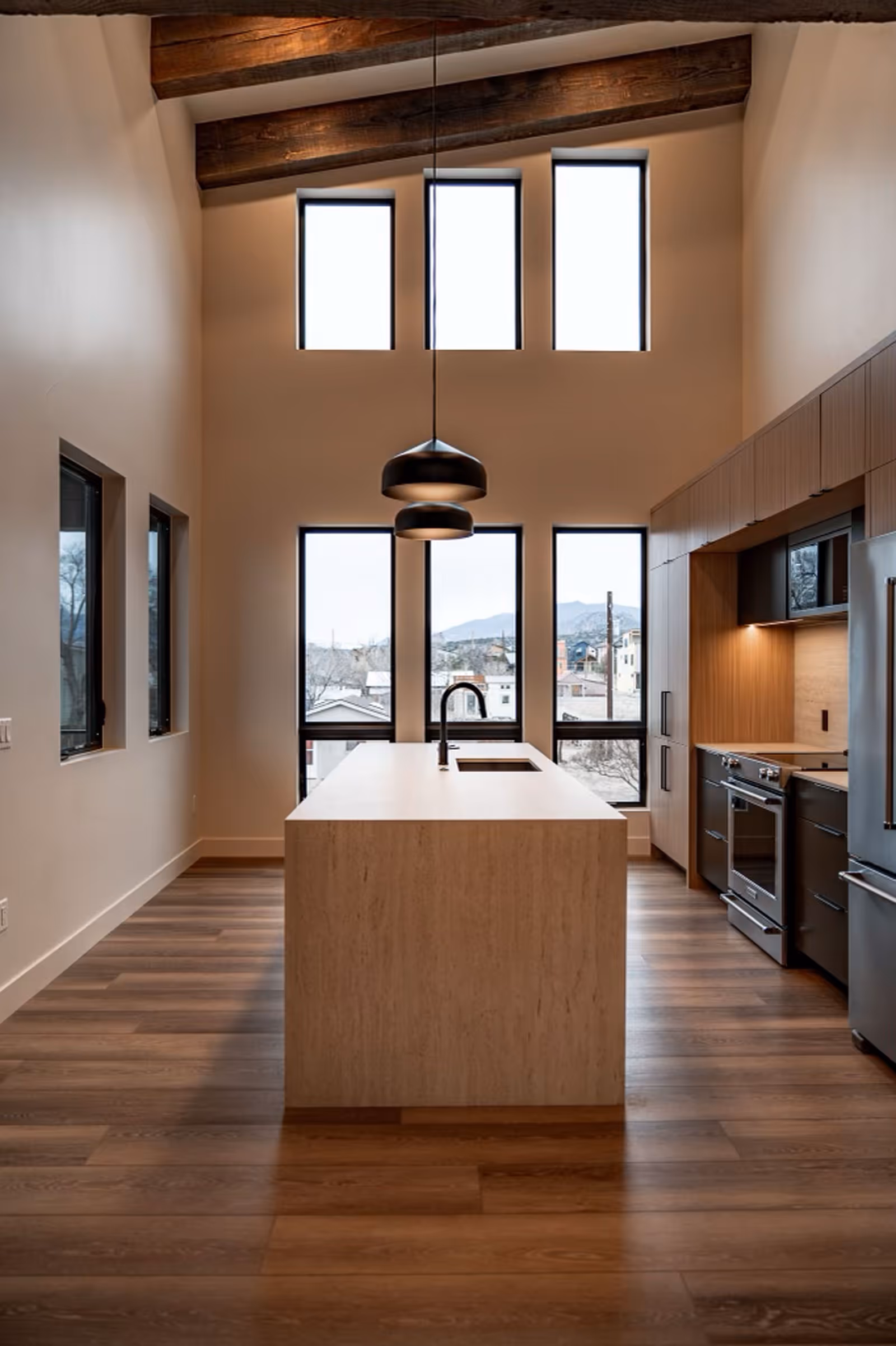 Modern open-concept kitchen with vaulted ceiling, exposed wood beams, center island, and large vertical windows overlooking a mountain town