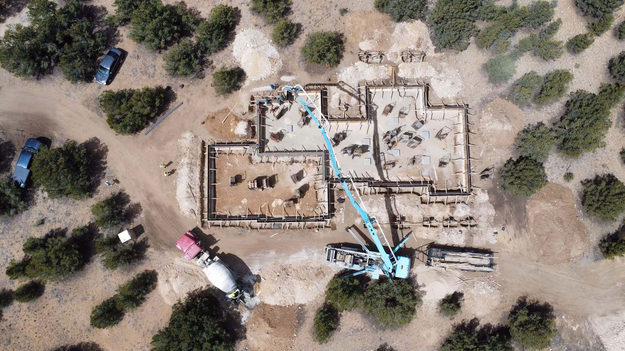 Top‑down drone shot of a house under construction on sandy ground dotted with scrubby trees, showing the concrete foundation and work vehicles.