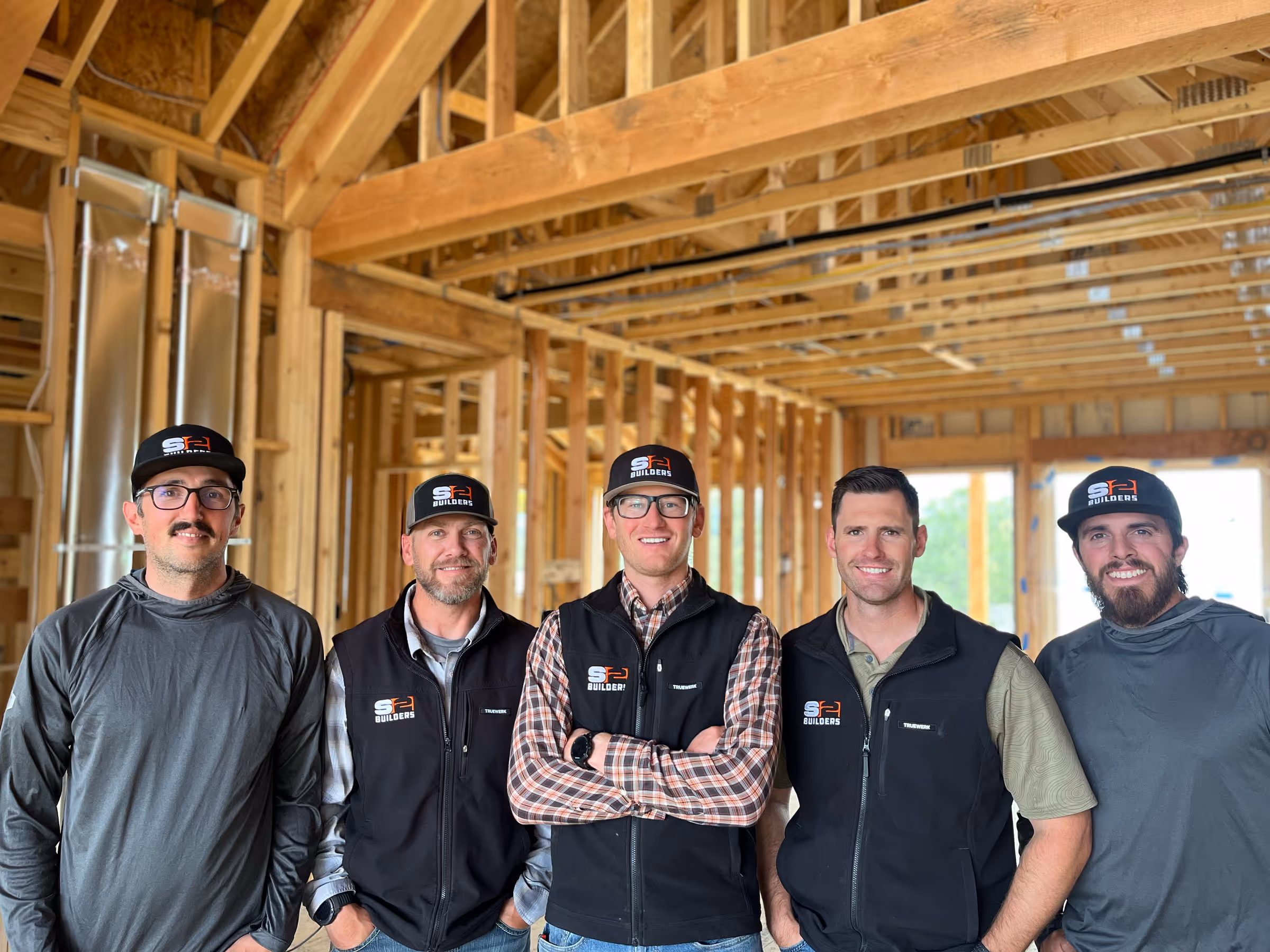 Five S2 Builders team members wearing branded hats and vests, standing inside a partially constructed wooden building frame.