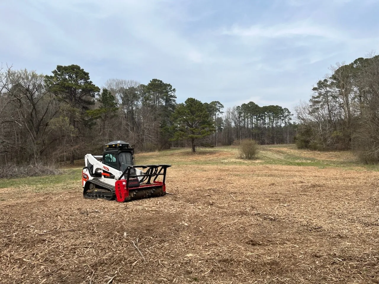 White and red skid steer mulching field with trees in background