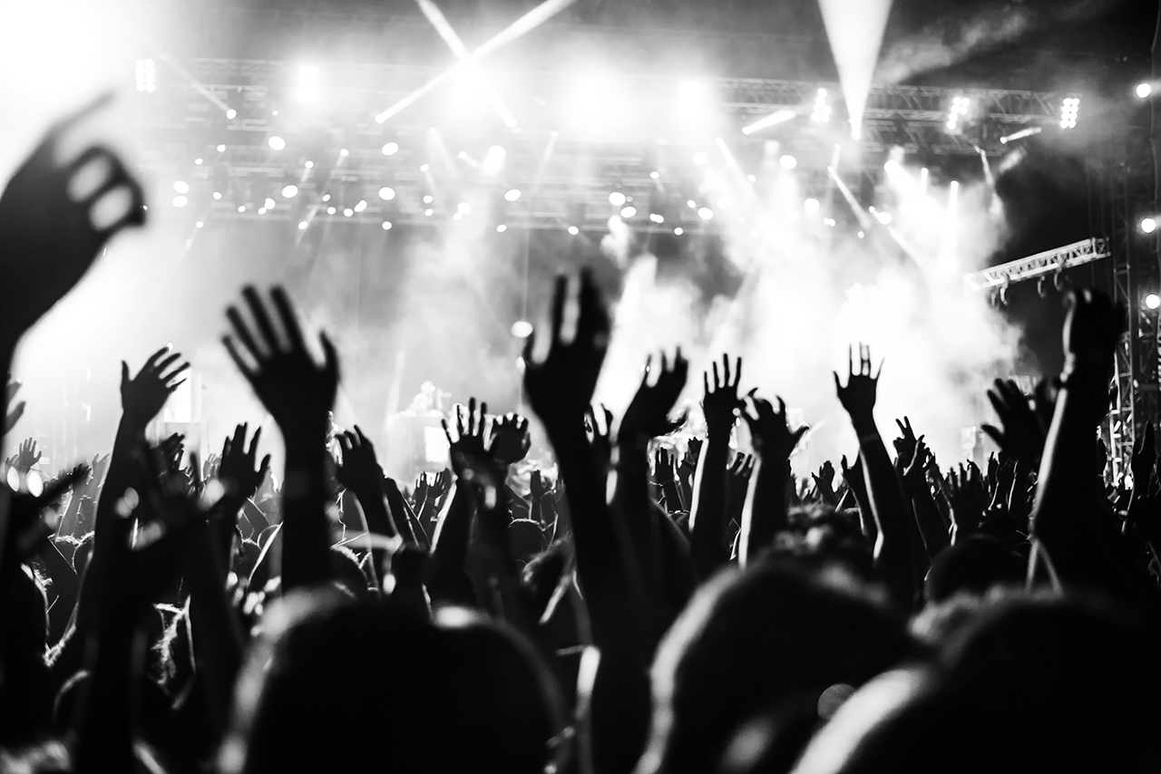Black and white photo of a large crowd at a concert, with many raised hands and stage lights shining in the background.