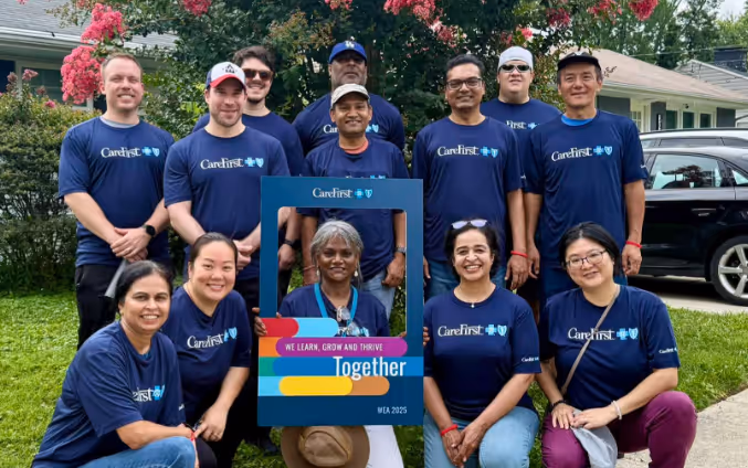 Group of diverse adults wearing matching blue CareFirst shirts posing outdoors with pink flowering trees in the background, holding a frame that reads 'We Learn, Grow and Thrive Together.'