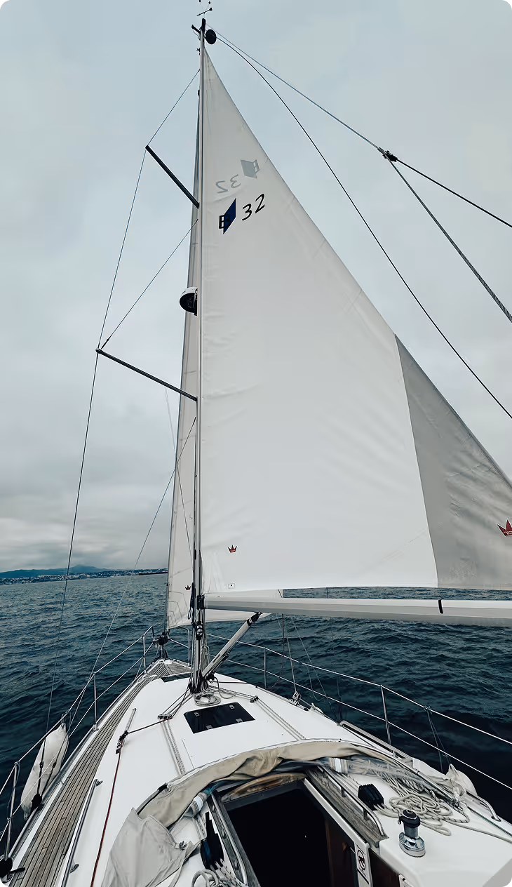 View from the bow of a sailing yacht with full sails, gliding through calm waters under a cloudy sky off the coast of Cascais.