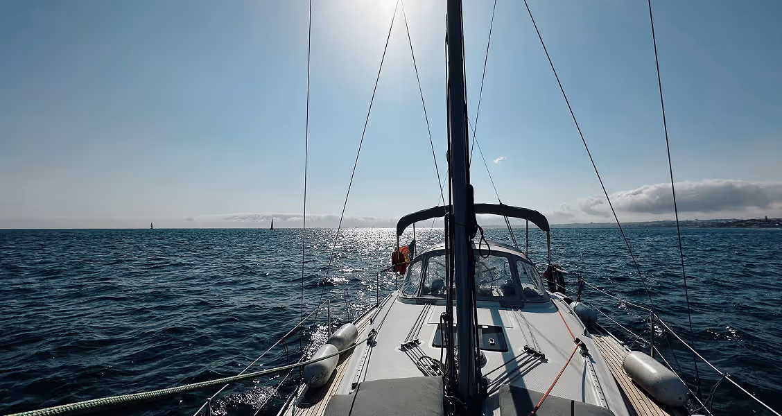 Forward view from the deck of a sailboat cruising along the Portuguese coast, with sunlit water and distant shoreline under a bright sky.