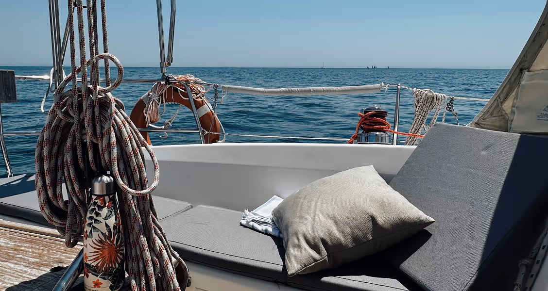 Cozy seating area on a sailboat with cushions and nautical ropes, overlooking the open sea on a clear, sunny day.