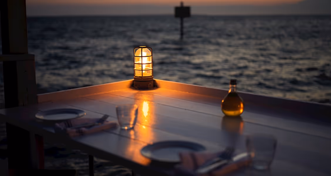 Romantic dinner table by the sea with warm lantern light at sunset, creating an intimate atmosphere.