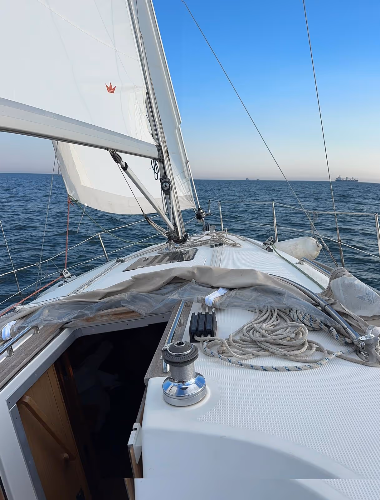 View from the deck of a sailing yacht gliding over calm ocean waters, with white sails unfurled and a clear blue sky above.