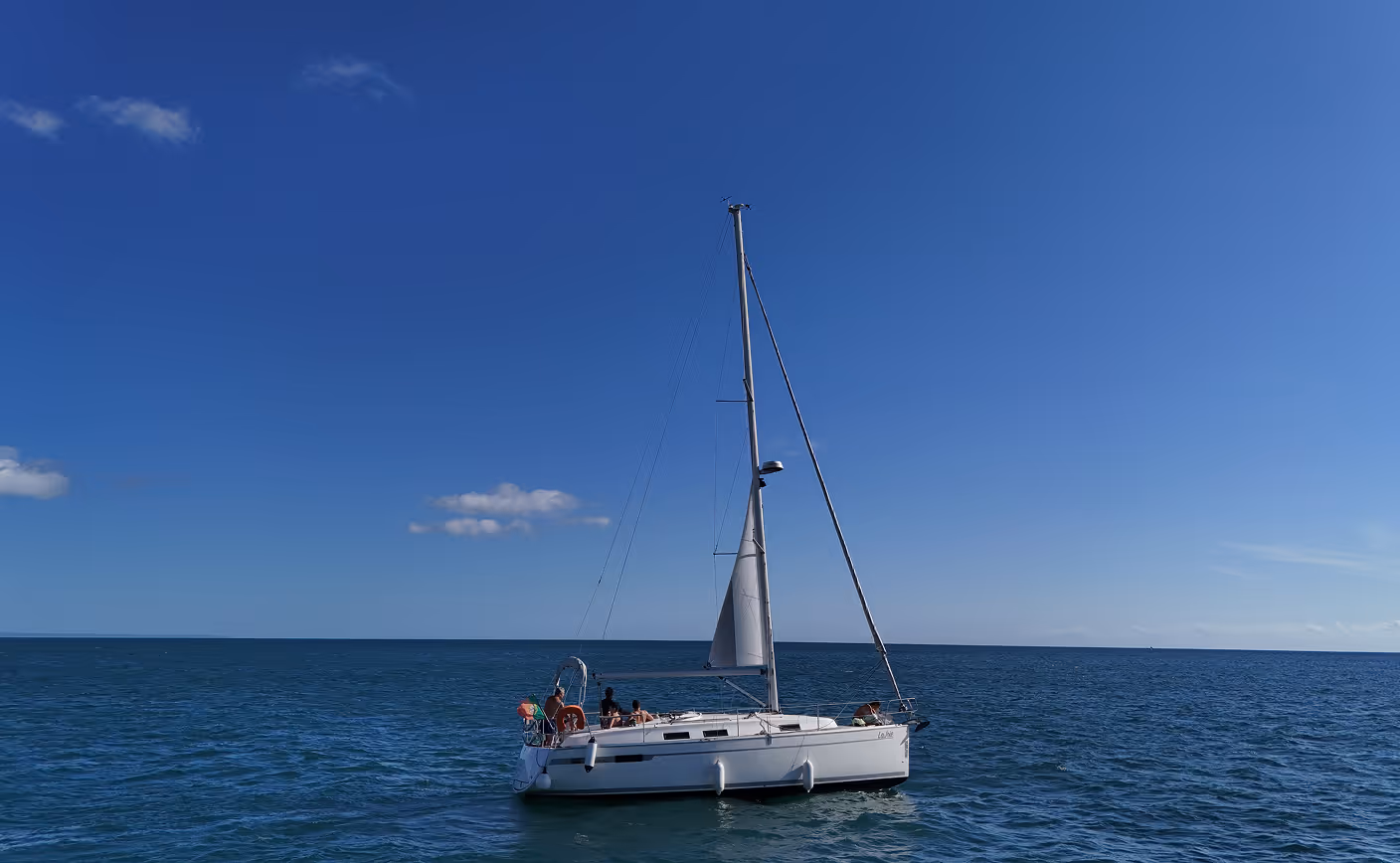 Close-up of a white sailboat with passengers relaxing on deck, sailing on the Atlantic Ocean under a bright blue sky.