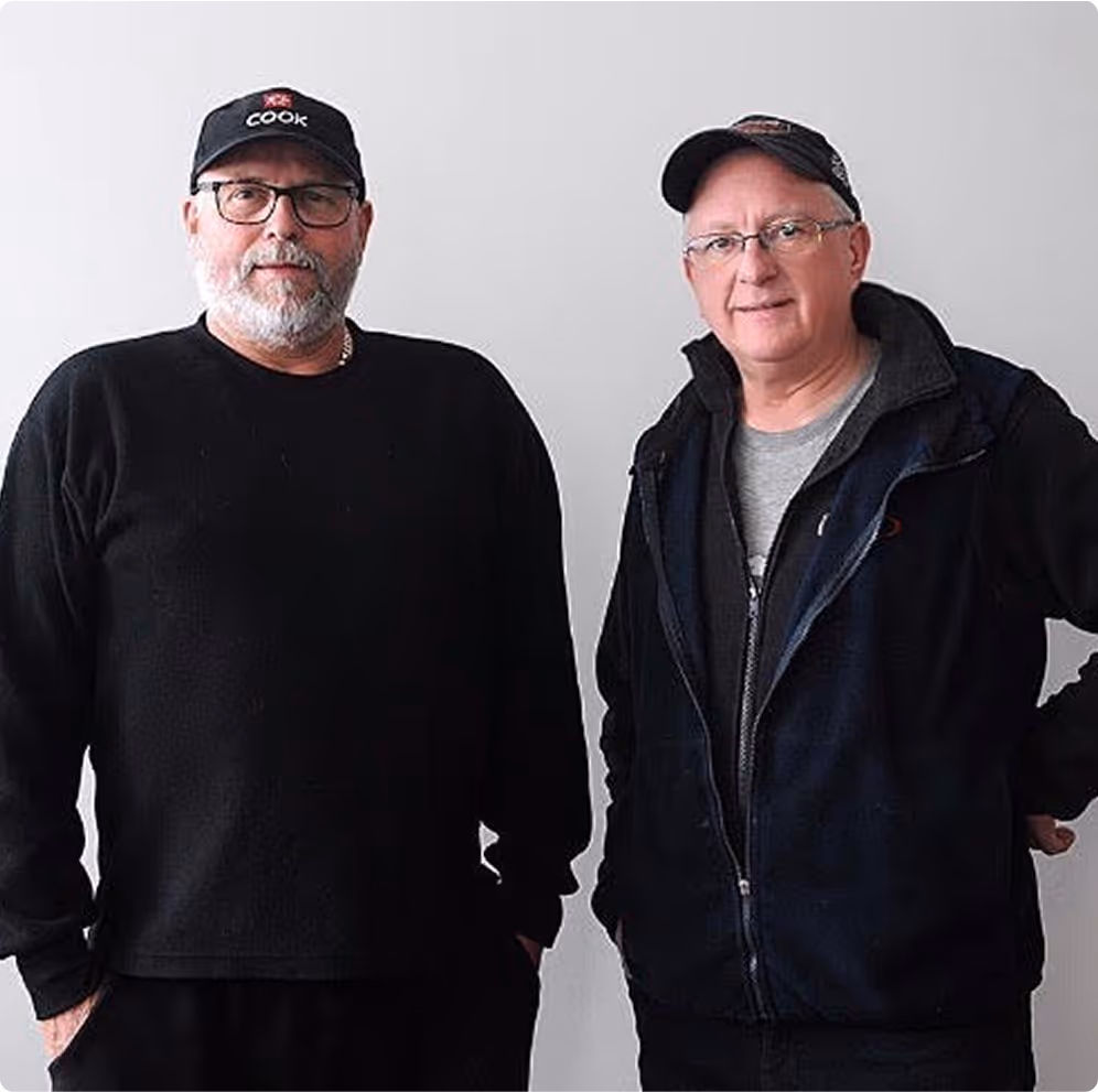 Two middle-aged men wearing glasses and dark casual clothing standing against a plain light background.