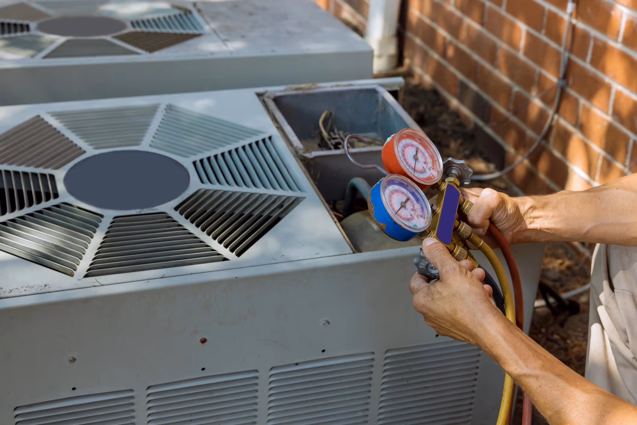 A technician adjusts pressure gauges on an outdoor air conditioning unit next to a red brick wall. 