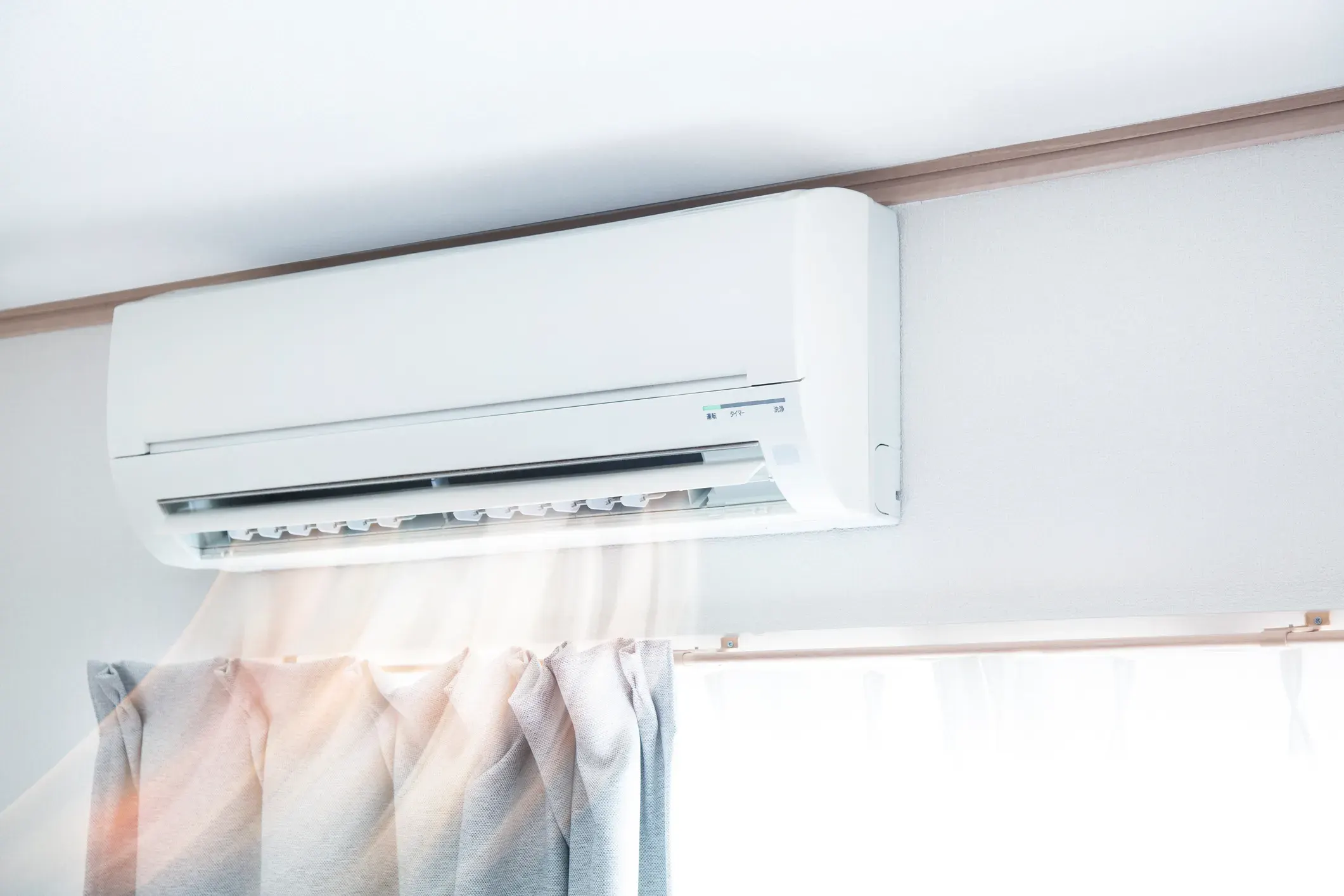 An indoor mini-split air conditioner blowing air, with curtains below a large window.