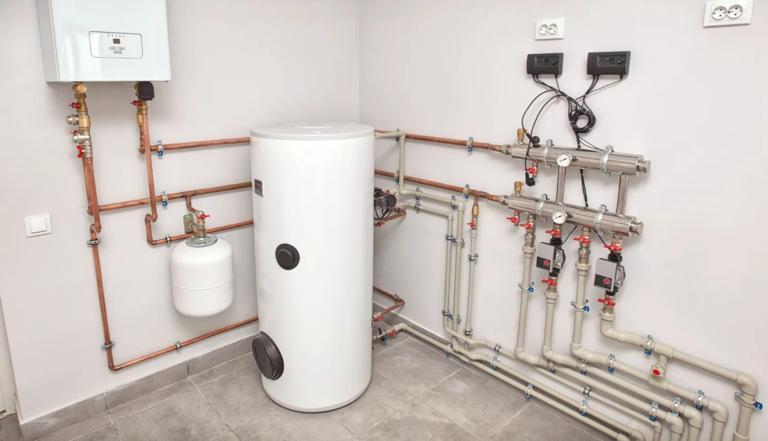 A complex boiler installation featuring a white storage tank, expansion vessel, and manifold with multiple circulation pumps and copper piping.