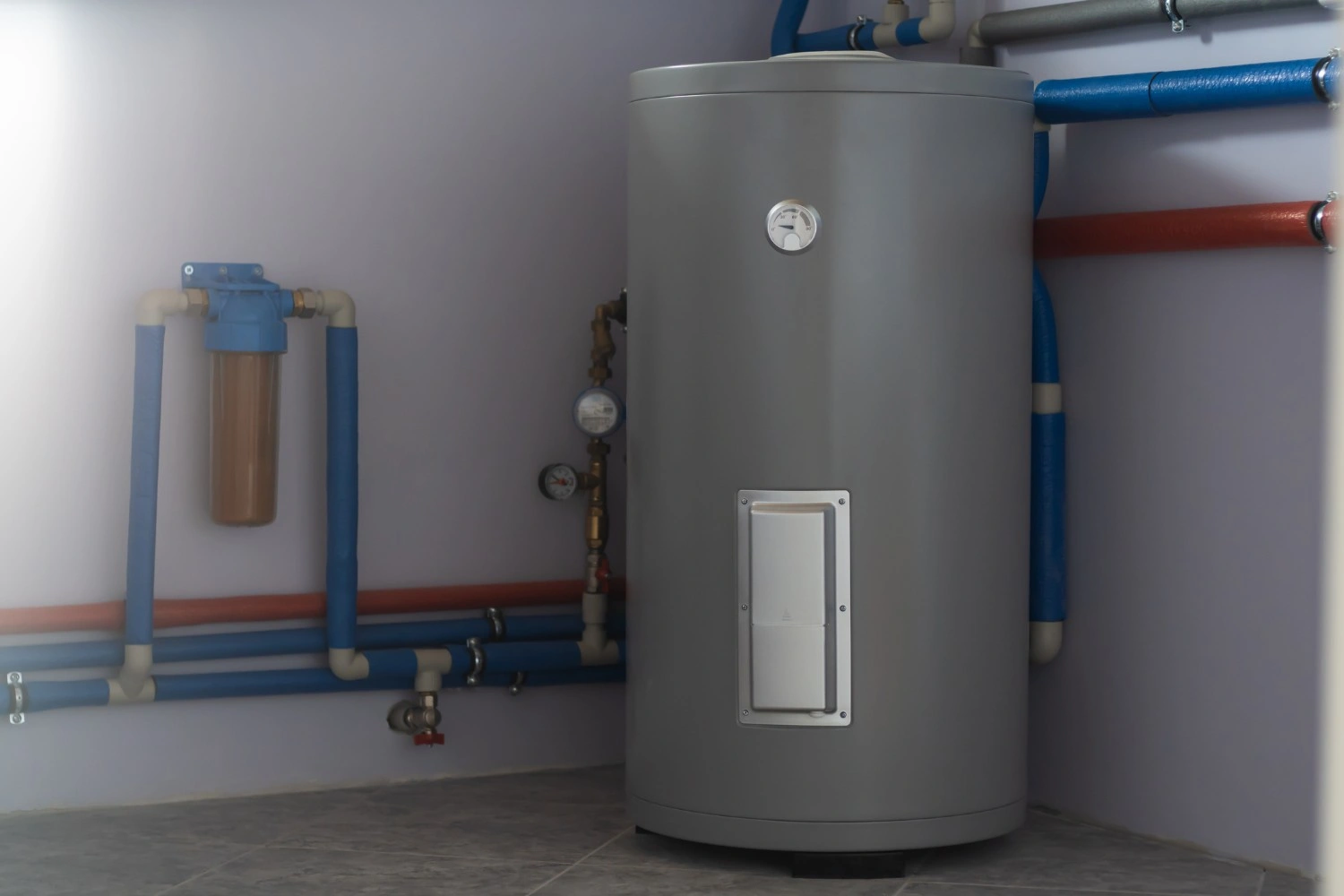 A grey cylindrical boiler tank is installed in a mechanical room with a water filtration system and insulated blue and red piping.
