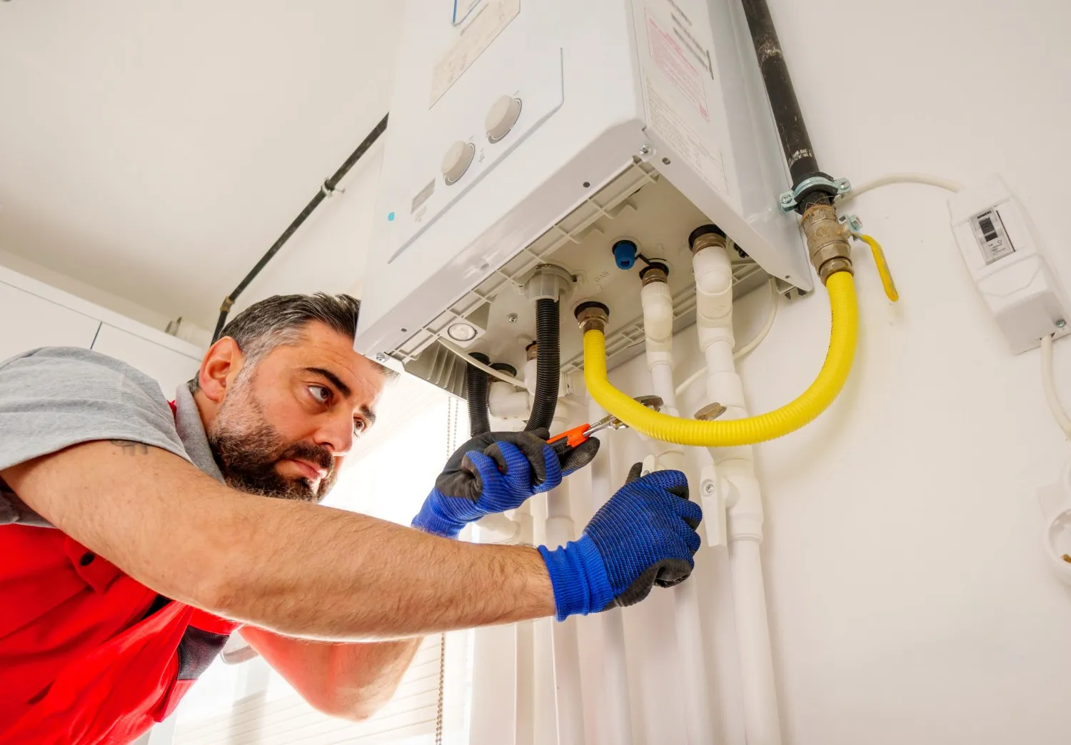 A technician in a red uniform repairs a wall-mounted boiler using a wrench on the unit's internal components.