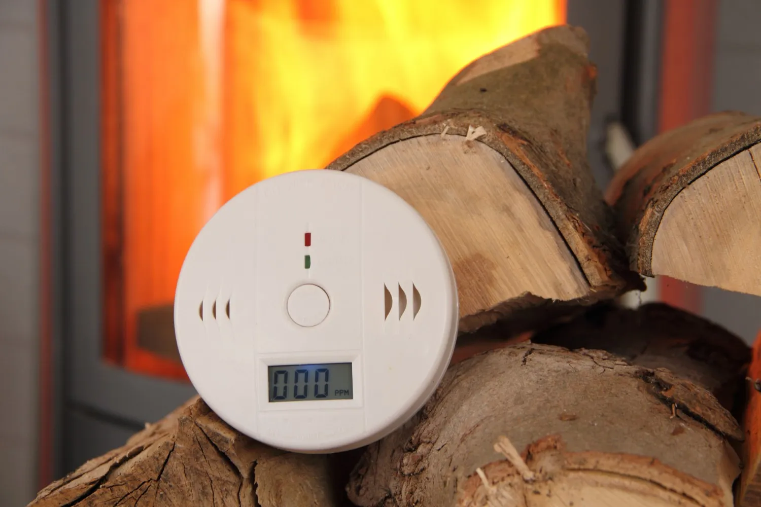 Digital carbon monoxide detector on firewood stack shows 000 PPM in front of active fireplace.
