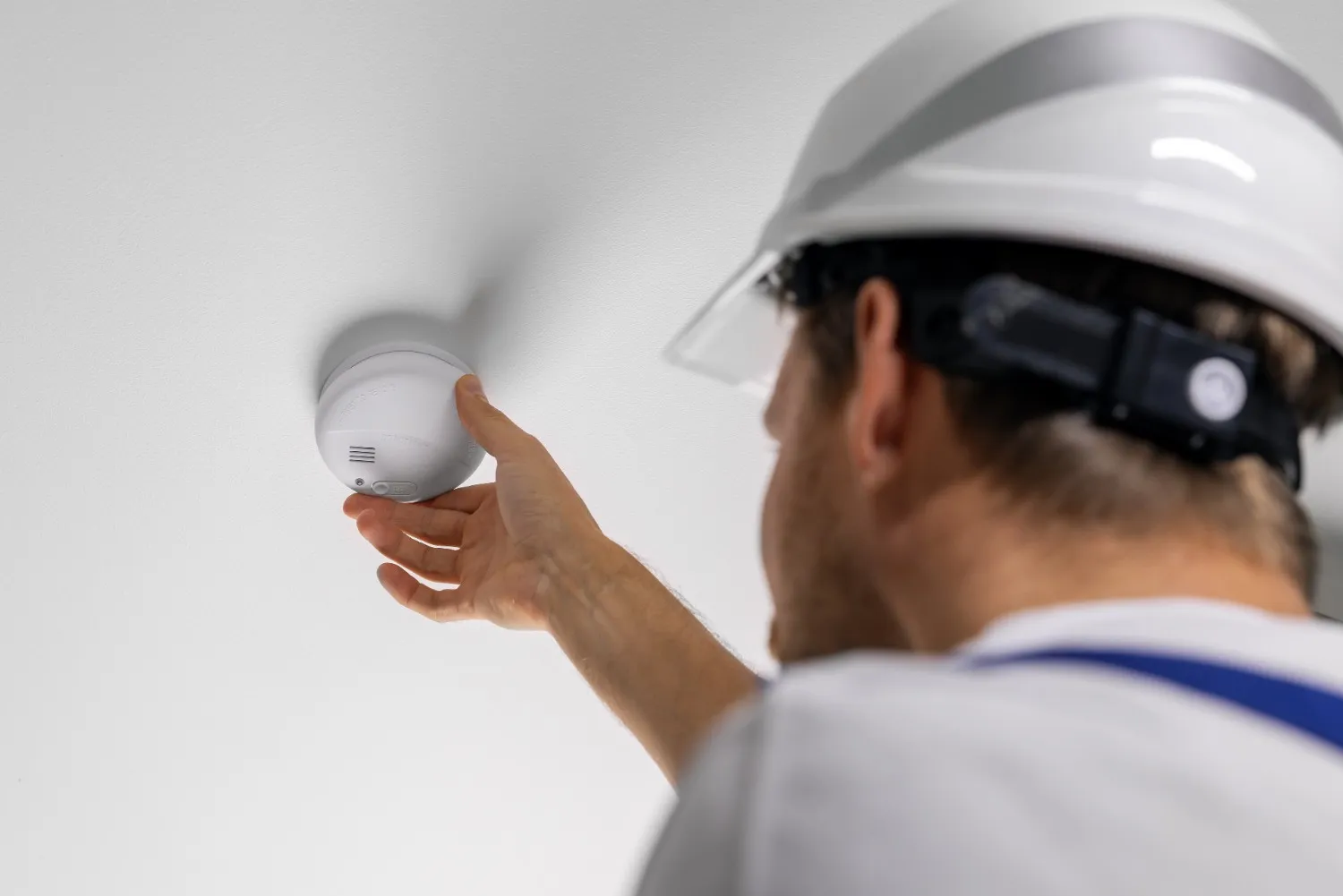 Person in white helmet adjusts ceiling-mounted smoke detector during safety inspection in residential setting.