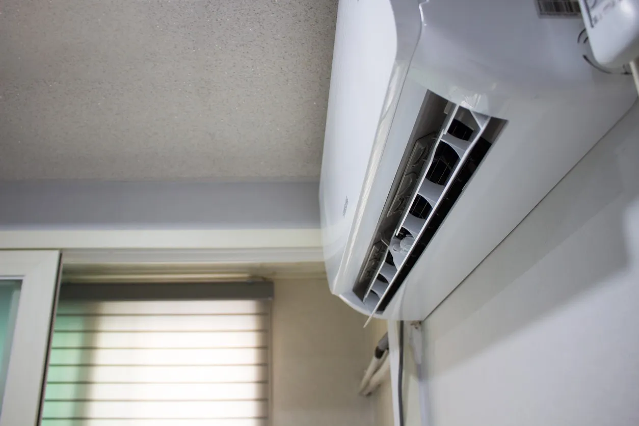 Air conditioning unit mounted on a wall, with vents facing down, next to a window with blinds partially closed.