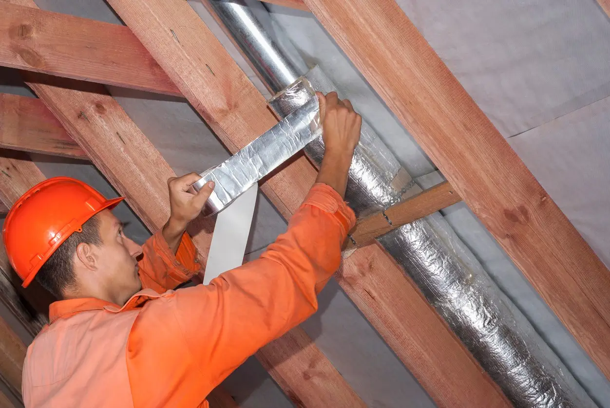 An HVAC technician in an orange hard hat and jumpsuit uses silver foil tape to seal the insulation around a metal duct pipe nestled between wooden roof rafters in an attic.