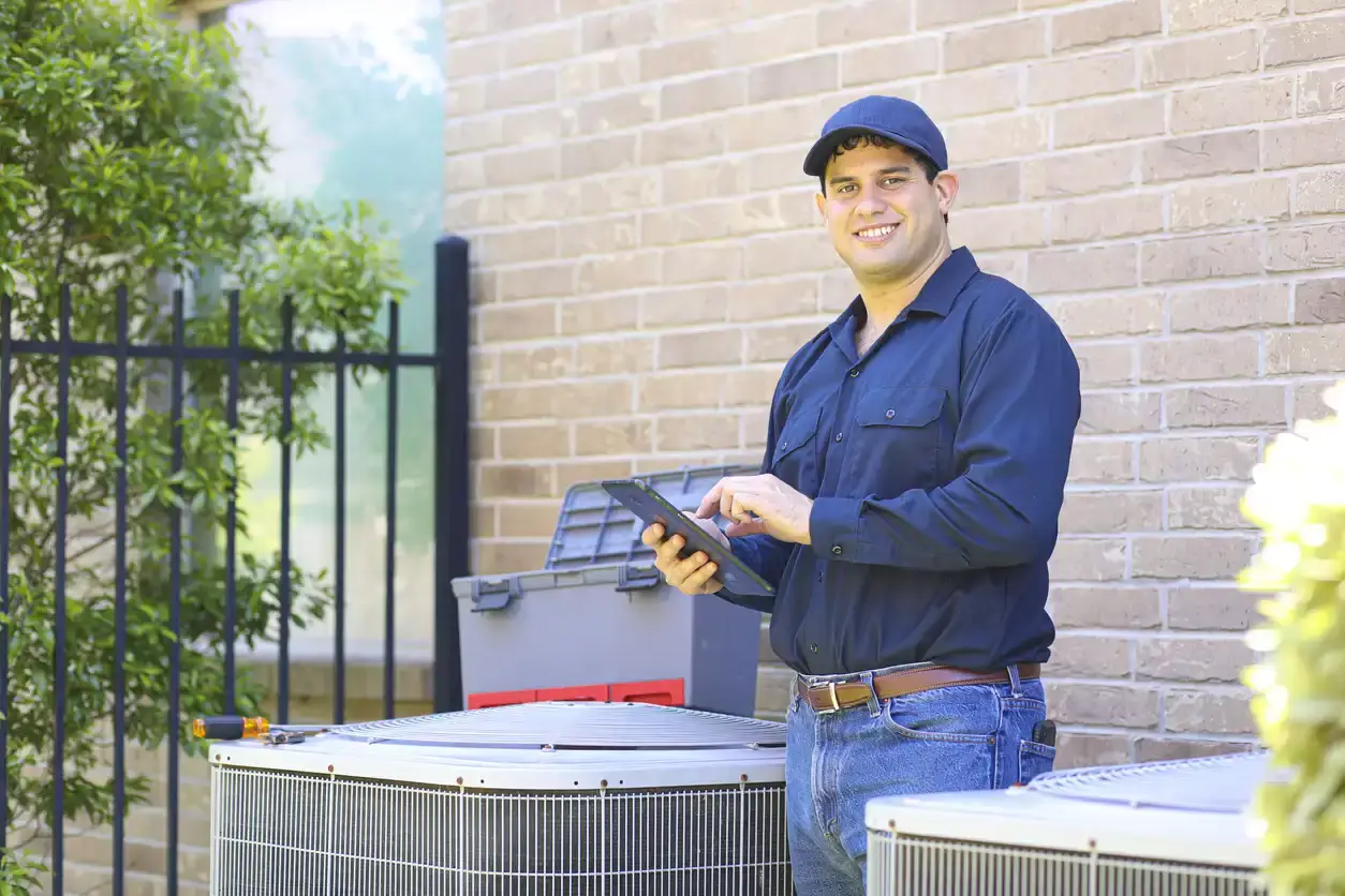 A smiling HVAC technician in a dark blue shirt, cap, and jeans stands outdoors next to a white heat pump unit while using a digital tablet for diagnostics and performance monitoring.