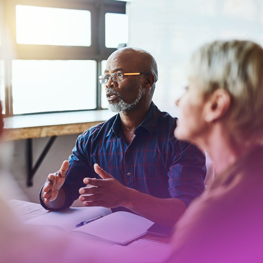 Man with glasses and beard speaking and gesturing with hands during a discussion in a bright room.