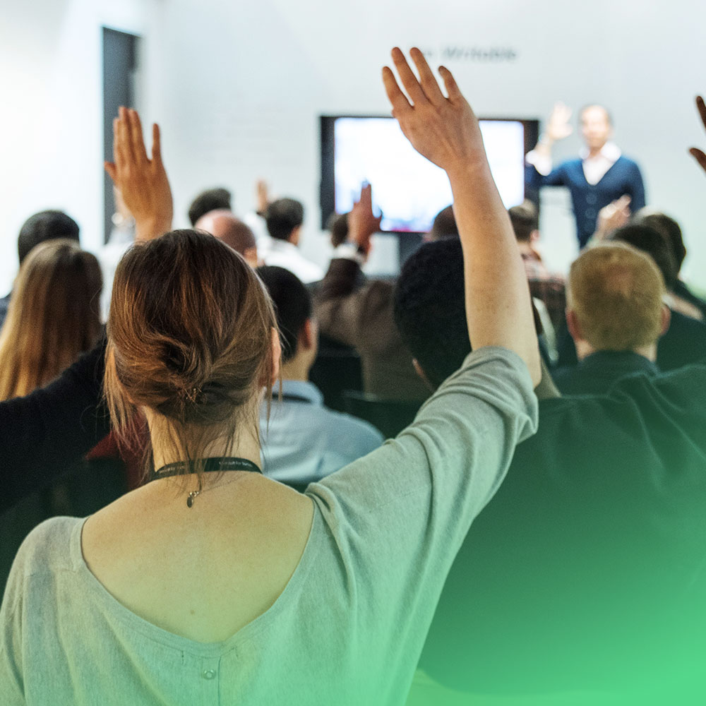 Audience members raising their hands during a lecture or workshop with a speaker in front.