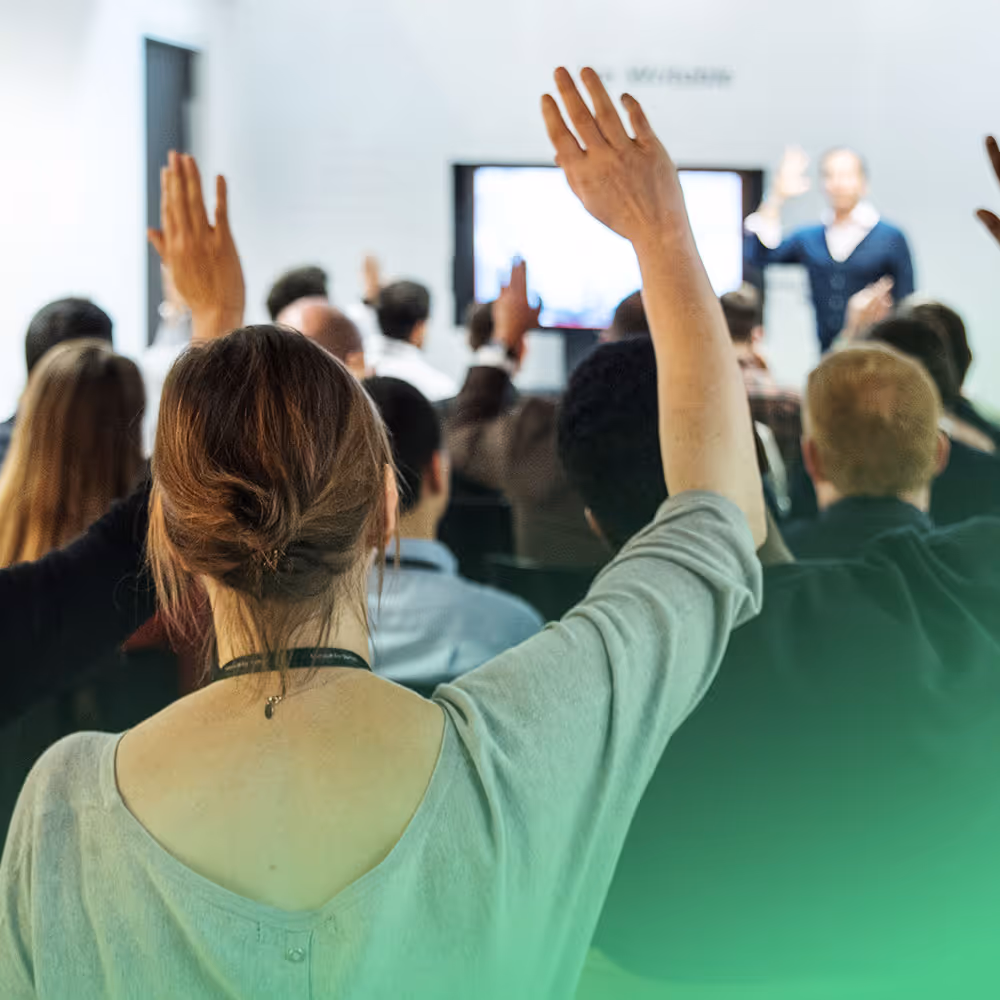 Audience members raising their hands during a lecture or workshop with a speaker in front.