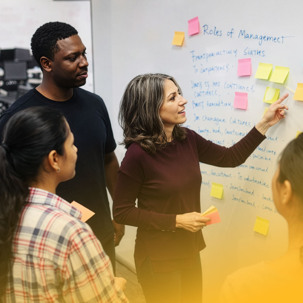 A woman pointing to colorful sticky notes on a whiteboard while three colleagues watch during a brainstorming session.