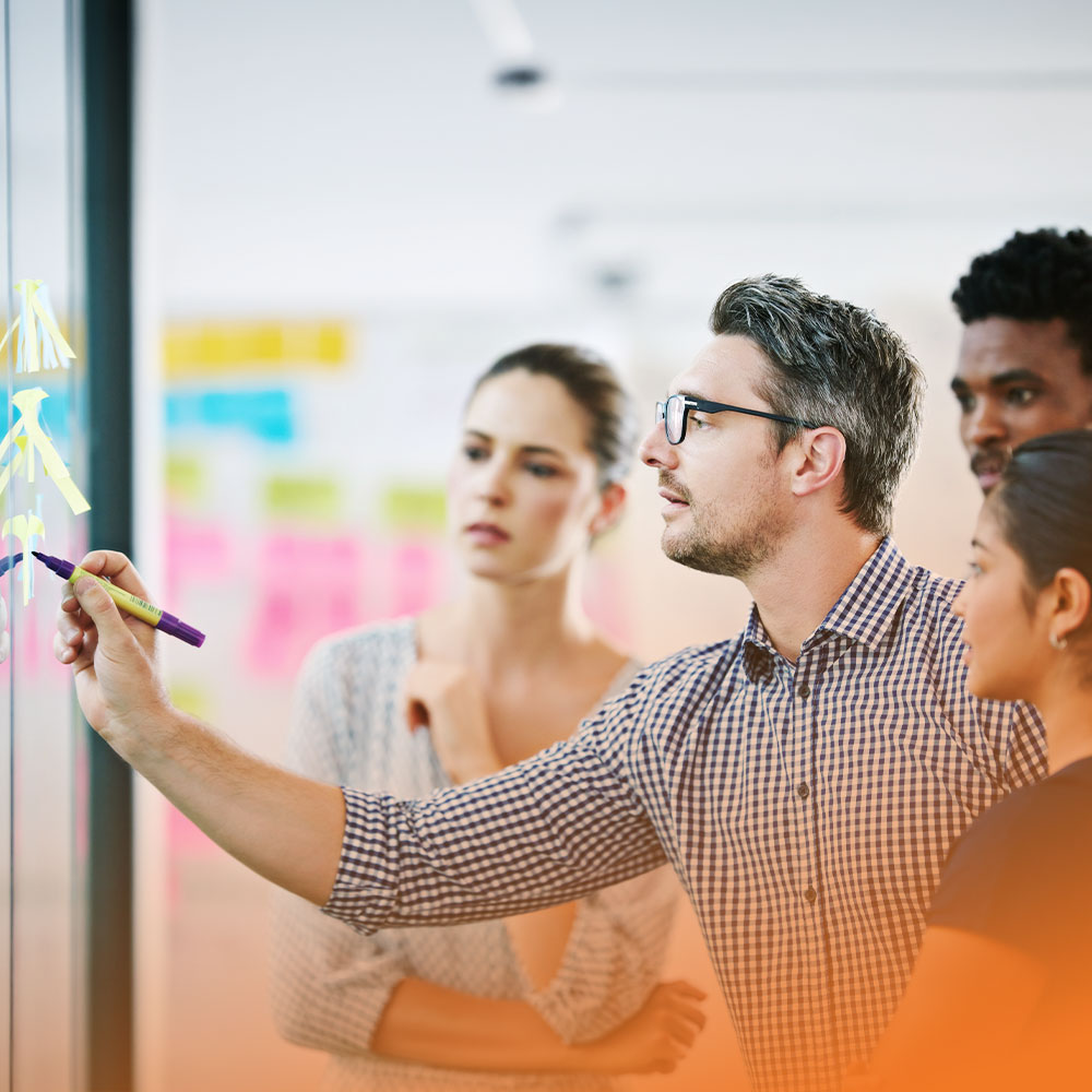 Four colleagues collaborating, with one man writing on a glass wall covered in colorful sticky notes.