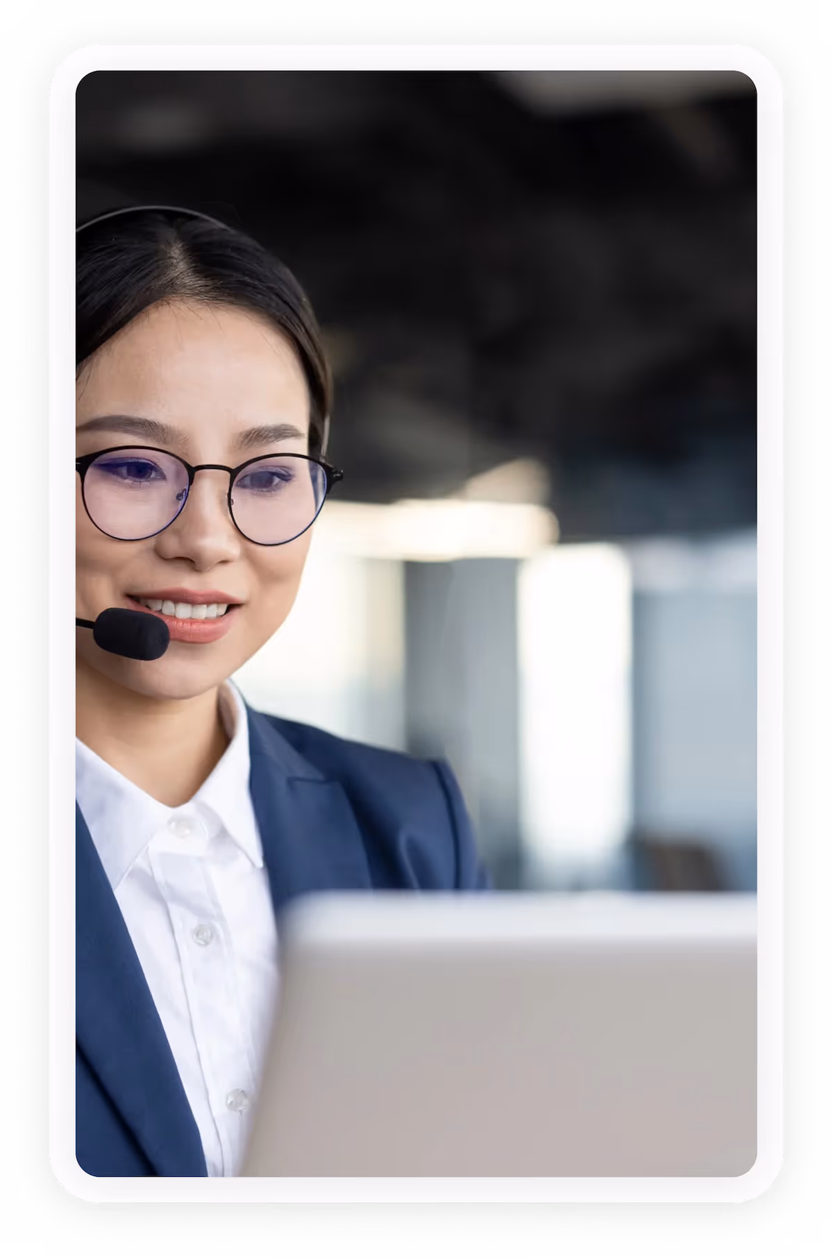 Smiling woman in glasses and headset working on a laptop in a bright office.
