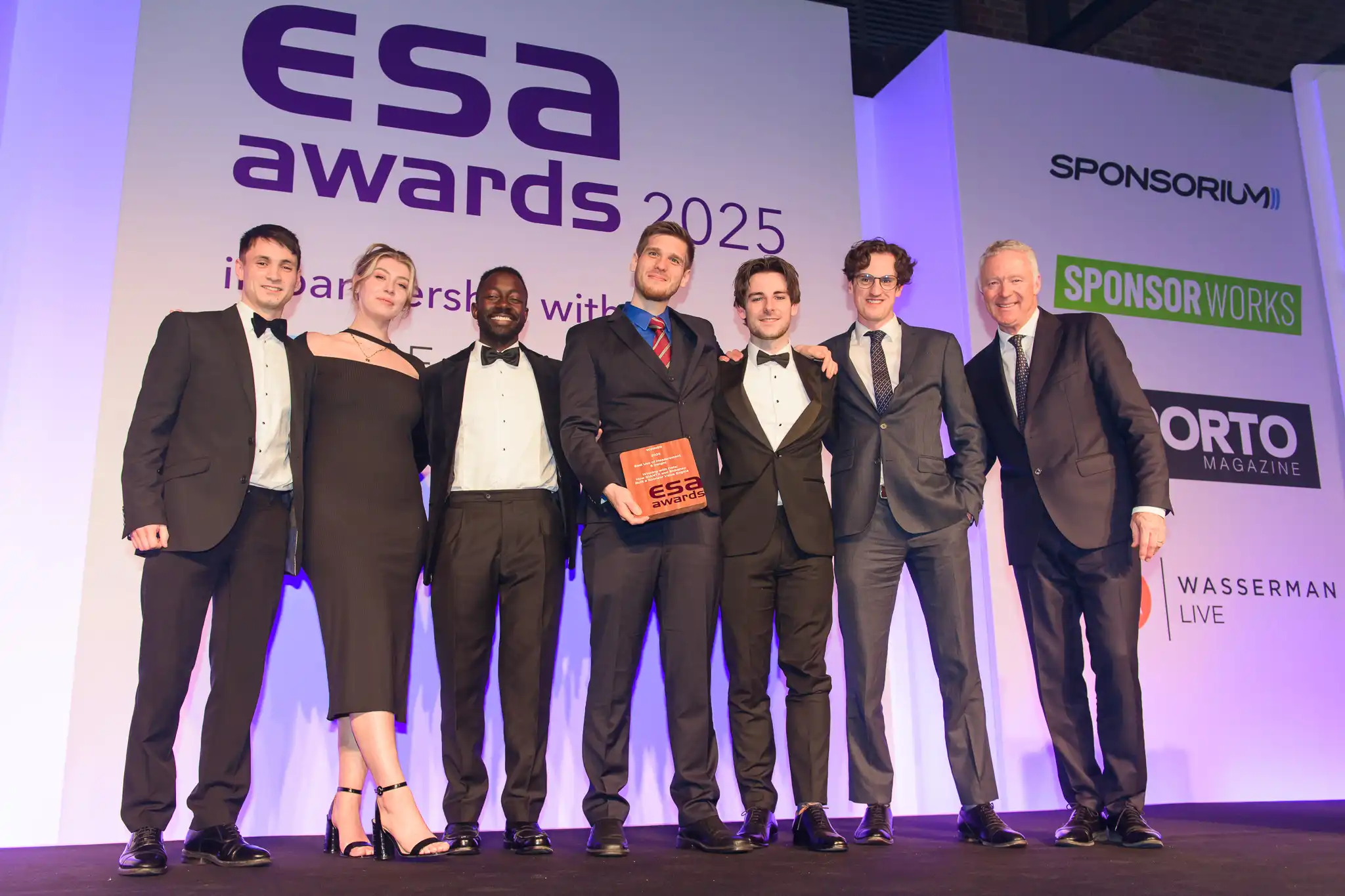 Shikenso with partner GIANTX in formal attire standing on stage at the ESA Awards 2025 ceremony, posing with an award plaque in front of a backdrop displaying event sponsors and logos.