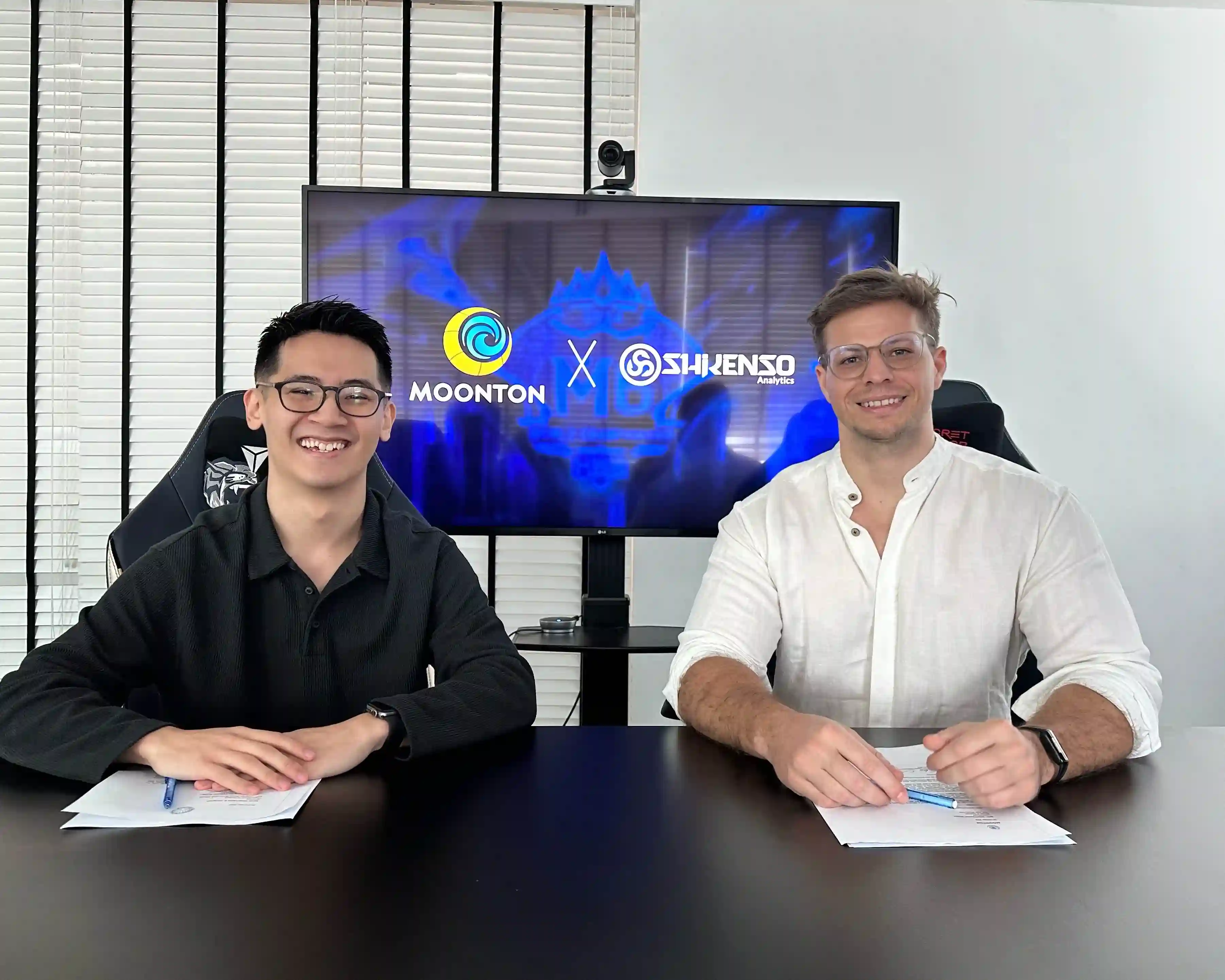 Two representatives from Moonton and Shikenso smiling and seated at a table with signed documents, with a screen behind them displaying the Moonton and Shikenso logos to announce their partnership.