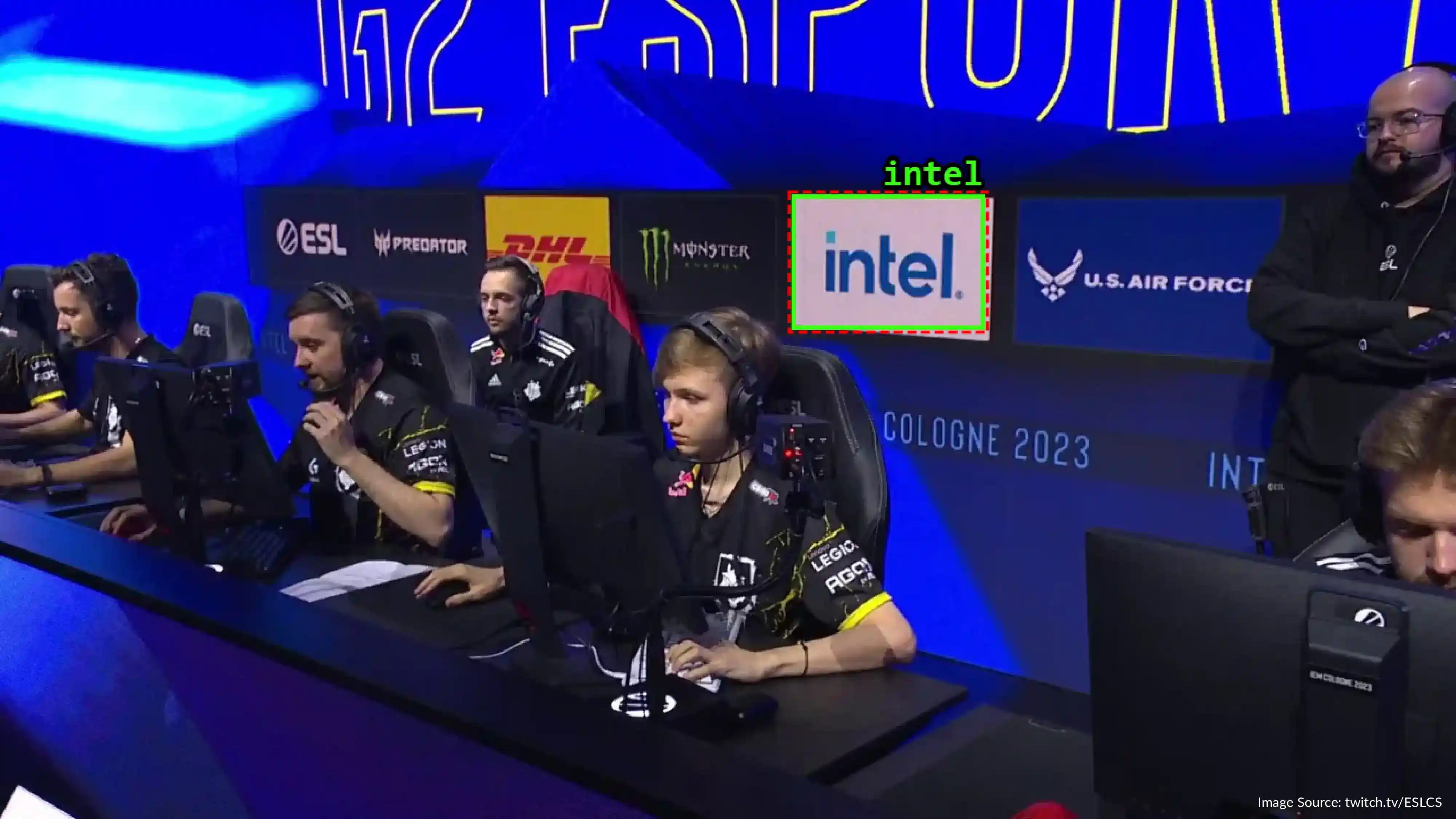 Esports team competing on stage at IEM Cologne 2023, sitting at gaming PCs with headsets, with sponsors including Intel, Monster, DHL, Predator, and U.S. Air Force displayed on the background wall.